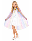 A young girl with blonde hair stands against a white background wearing a white dress and a pastel rainbow unicorn cape with blue pom pom tie.
