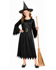 A young girl modeling a black witch costume dress with a pointed hat and holding a broomstick against a plain white background.