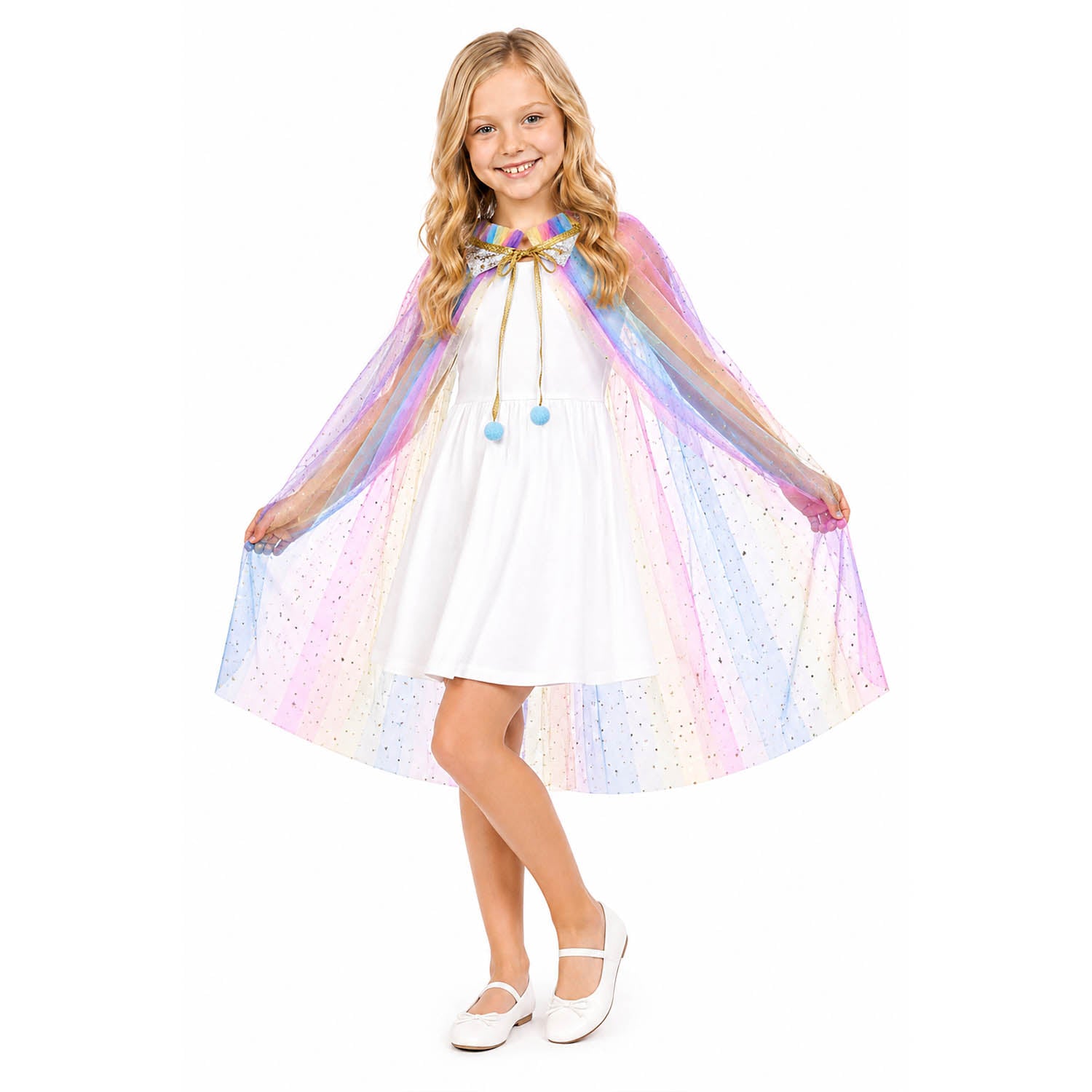 A young girl with blonde hair stands against a white background wearing a white dress and a pastel rainbow unicorn cape with blue pom pom tie.