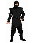 A child modeling a black ninja costume with a hooded mask, a large shoulder cape, loose pants, and black boots, holding a toy sword against a plain white background.