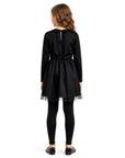 A young girl viewed from the back wearing a black long-sleeve dress with a tulle hem, black tights, and black flats, set against a plain white background.
