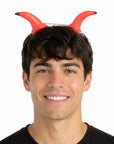 A smiling man wearing a pair of red devil horns with black tips on a white background.