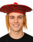 A man wearing a red and black tartan plaid bonnet hat with a small red pom-pom on top, set against a plain white background.